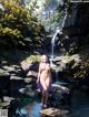 A naked woman standing in a river next to a waterfall.