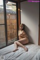 A naked woman sitting on a bed in front of a window.